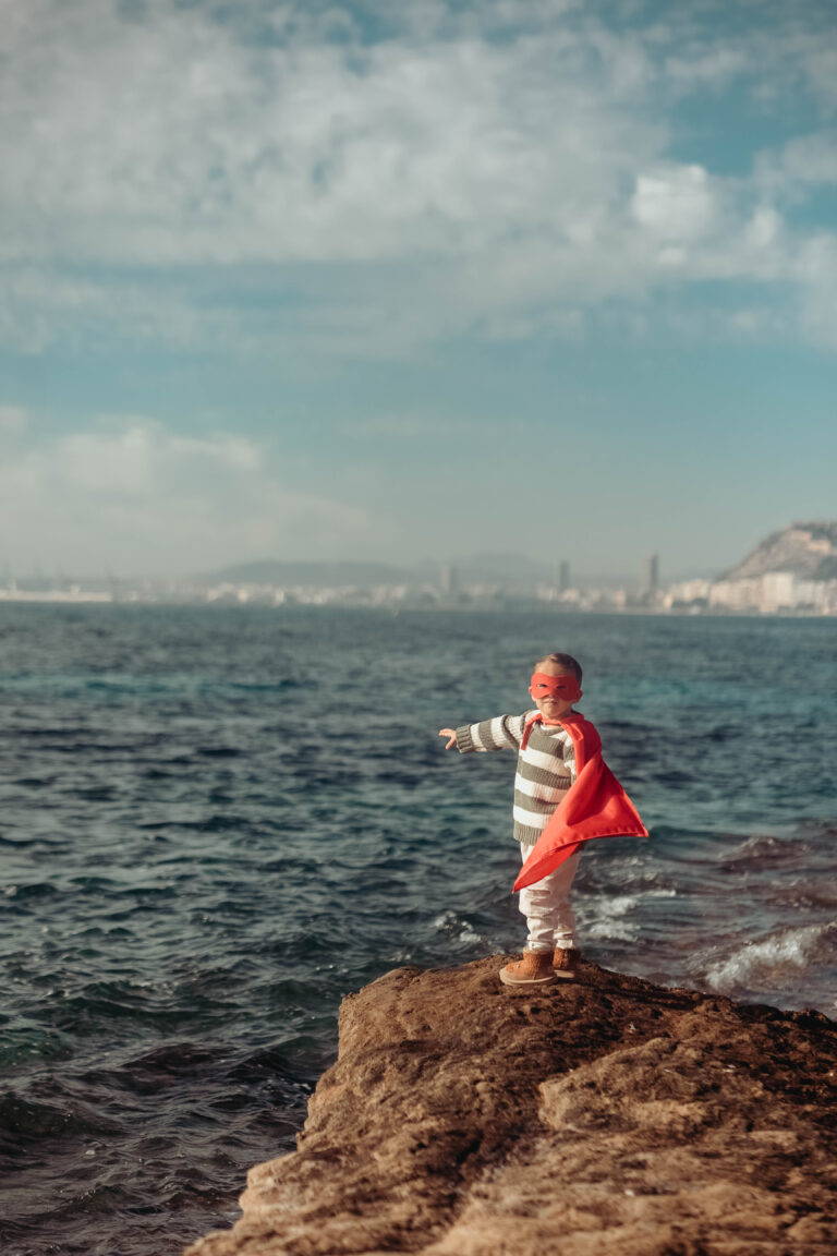 Pequeño superhéroe posando frente al Mediterráneo en Alicante, con una gran sonrisa y espíritu de aventura.